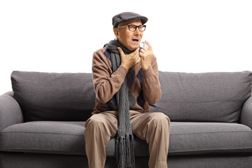 Obraz premium Elderly gentleman with a sore throat sitting on a sofa and spraying a medication