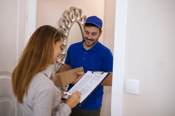 Woman Signing For Package From Courier At Home. Smiling young delivery man holding a cardboard box while beautiful young woman putting signature in clipboard