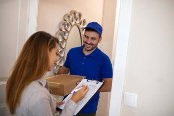 Woman Signing For Package From Courier At Home. Smiling young delivery man holding a cardboard box while beautiful young woman putting signature in clipboard