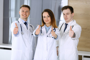 Fototapeta premium Group of modern doctors standing as a team with thumbs up or Ok sign in hospital office. Physicians ready to examine and help patients. Medical help, insurance in health care, best treatment and