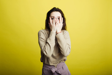 An image of a happy young girl standing isolated on a yellow background covered her face with her hands