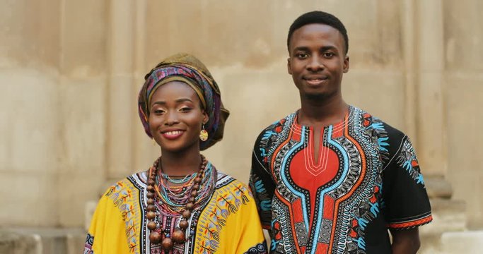 Portrait shot of the attractive young African couple in the colorful traditional clothes standing together at the wall and smiling happily to the camera. Outdoors.