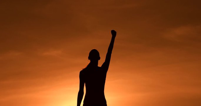 Strong, victorious , and motivated young woman raising her fist up in the air against sunset sky. People determination and overcoming adversity concept.