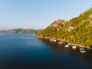 Obraz premium Aerial drone perspective of luxury Turkish gulets and yachts in the deep blue and turquoise waters of the mediterranean sea. The bay and cliffs protect the boats from the strong winds.