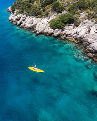 A lone girl paddling a yellow kayak in remote mediterranean waters, exploring the Turkish coastline. Beautiful blue water and coastline with amazing visibility. Shot aerially with a drone.