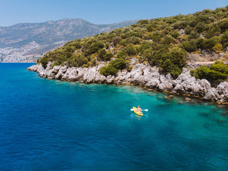 Fototapeta premium A lone girl paddling a yellow kayak in remote mediterranean waters, exploring the Turkish coastline. Beautiful blue water and coastline with amazing visibility. Shot aerially with a drone.