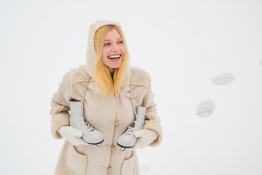 Young blond woman in sweater and funny hat holding white skates over shoulder in freezing winter day. Beautiful woman walking in winter park and feeling wonderful.