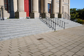 The steps and columns of a large government courthouse building with a red door. The steps have a black handrail in the center. There's a brick pathway to the large marble building.