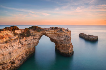 Algarve beach, romantic couple watching the sunset. Loving moment under natural arch carved in stone is a tourist attraction of the south coast of Portugal. Panoramic view from the cliff.