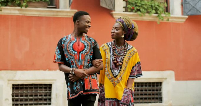 Portrait shot of the African young romantic good looking man and woman in stylish traditional clothes standing hand in hand on the street and looking at each other with lve and smiles.