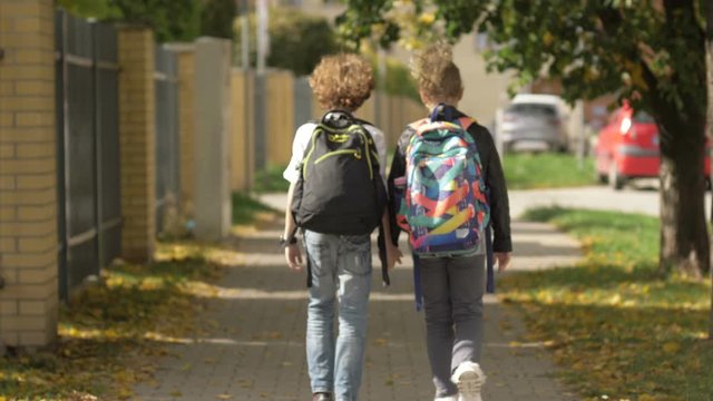 Two Child Girl and Boy Walking on city streets past cars. Sister with her Brother Back to School. Sun is on Background. Back View