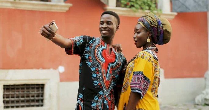 African young stylish and attractive couple in traditional outfits standing together outside and posing to the smartphone camera while taking selfie photo.