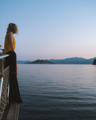Fototapeta premium Beautiful blonde girl leaning over the edge of a yacht looking at sunset over the mountains in the distant background. 