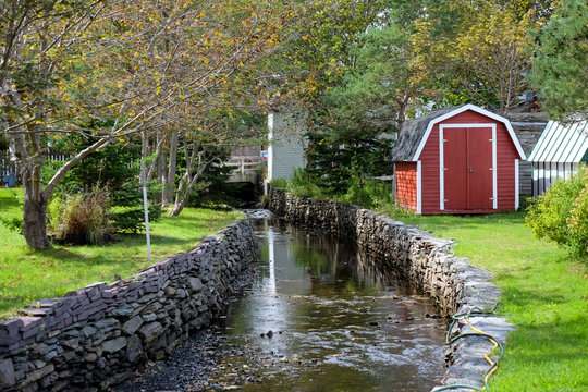 A small red building, shed or storage structure with white trim. A small stream with slow running water and rock walls on the left of the property. Green grass and trees in the foreground.
