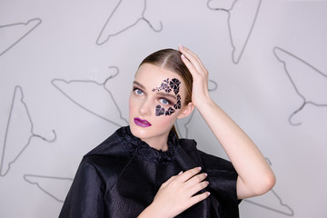Fashion portrait of a girl with clean skin, bright professional make-up, purple lipstick, face with black ivy tattoo around her eyes with rhinestones. Gray background, pattern of hangers. smoky eyes