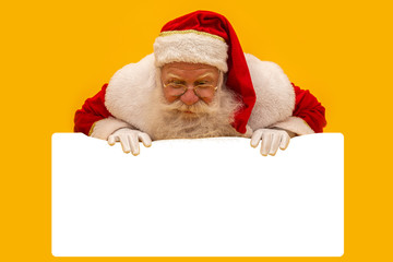 Happy Santa Claus looking out from behind the blank sign isolated on yellow background with copy space