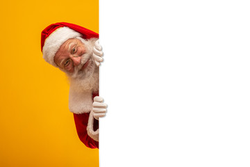 Happy Santa Claus looking out from behind the blank sign isolated on yellow background with copy space