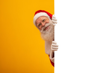 Happy Santa Claus looking out from behind the blank sign isolated on yellow background with copy space - obrazy, fototapety, plakaty