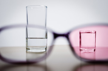 Optimism, illustrated by comparing less than half full glass in one frame of glasses with full smaller glass through rose tinted lens in another frame. Amount of water is about same in both glasses.