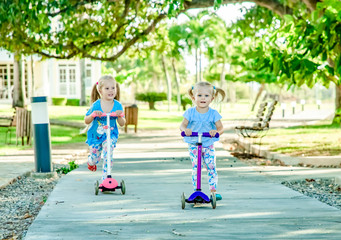 Two little girls, sisters riding scooters in a summer sunlit square under a big tree