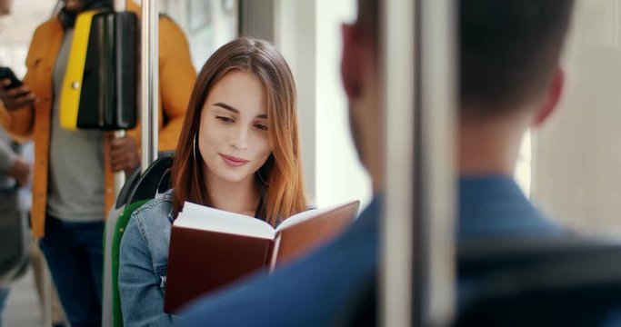 Caucasian young beautiful and happy girl reading a book in the tram and smiling, then flirting with a young handsome man who sitting in front of him. Rear.