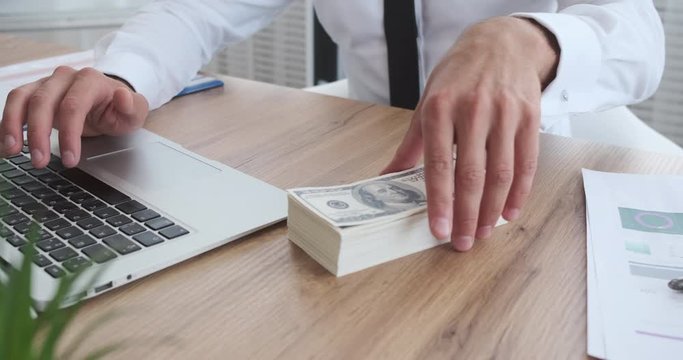 Businessman counting and smelling bribe money received while working on office laptop