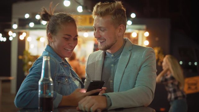 Beautiful Young Couple is Using a Smartphone while Sitting at a Table in an Outdoors Street Food Cafe. They're Browsing Internet or Social Media, Watching Videos. They are Happy and Smile.