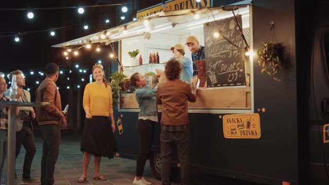 Food Truck Employee Hands Out Freshly Made Burgers, Fries and Drinks to Happy Young Hipster Customers Who Have a Party Outisede. Commercial Truck Selling Street Food in a Modern Cool Neighbourhood.