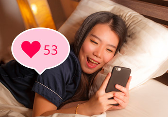 Young beautiful and happy Asian girl getting social media likes. Gorgeous Chinese woman in bed at night excited getting positive feedback on internet  app smiling cheerful
