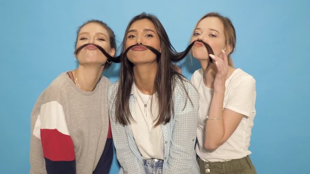 Three young beautiful smiling hipster girls in summer clothes. Sexy carefree women having fun.Positive models going crazy near blue wall in studio.They make mustache with hair.