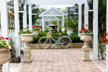 Fototapeta na wymiar Retro ladies bicycle with basket standing against garden retaining brick wall, underneath white wooden pergola with lush green shrubs and red flowers in background