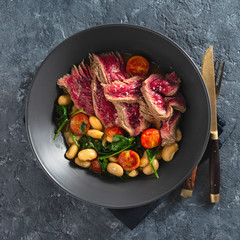 Dinner plate with beef steak with white beans, spinach and tomatoes on dark stone background top...