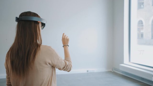 Young Hipster Woman in Holographic Augmented Reality Glasses Standing in Empty Office and Map it. She's Moving Virtual Pieces Around the Space. Room Has Tracking Points for Video Software.