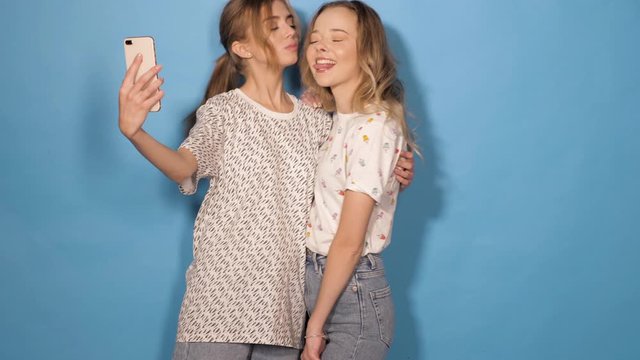 Two beautiful smiling girls.Women in summer hipster clothes taking selfie self portrait photos on smartphone. Models making funny faces and having fun in studio.They show tongue and kiss her friend