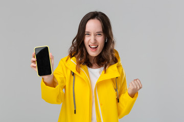 Smiling brunette woman in a yellow raincoat expressing emotion of success showing smartphone screen isolated over grey background. Concept of success.