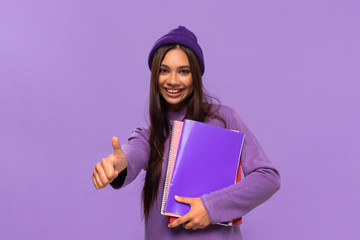 Smiling african-american tto teenager student in a hat and sweater holding folders and showing thumb up standing isolated over purple background. Concept of education