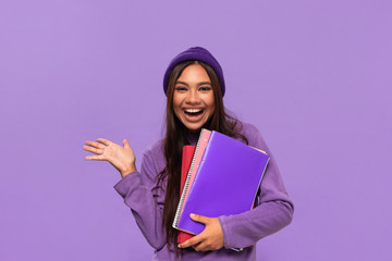 Pretty african-american teenager in a hat and sweater holding folders and expressing emotion of surprise standing isolated over purple background. Concept of education