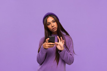 Smiling african-american girl in a hat and sweater holding smartphone isolated over purple background.