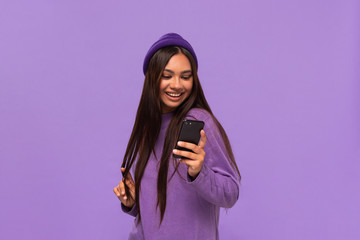 Smiling african-american girl in a hat and sweater holding smartphone isolated over purple background.