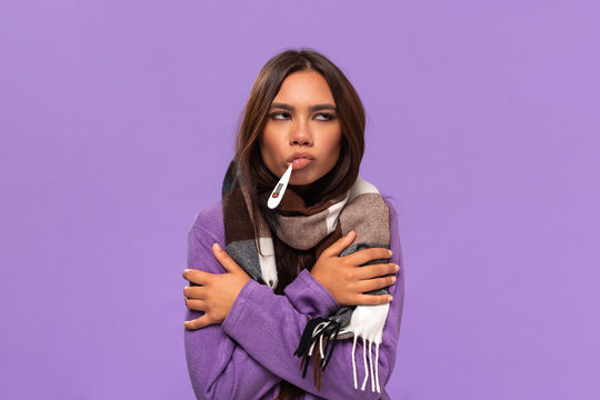 Pretty african-american girl in a sweater and cosy checked scarf measuring thermometer isolated over purple background. Catching flu in winter.