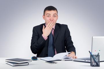 Young business man sitting at the office and looking shocked at the camera covering his mouth with his hand
