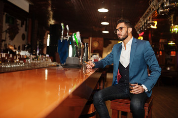 Handsome well-dressed arabian man with glass of whiskey and cigar posed at pub.