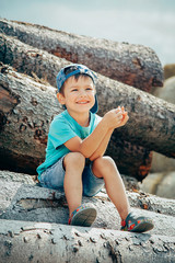 A little boy in a blue baseball cap and short shorts sits on a log and laughs merrily