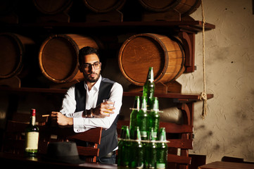 Handsome well-dressed arabian man with glass of whiskey and cigar posed at pub.