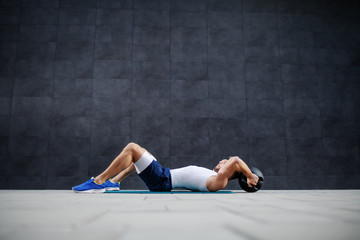 Side view of handsome muscular caucasian man is shorts and t-shirt lying down on mat and doing exercises with kettle bell.