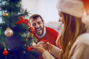 Laughing handsome bearded caucasian man helping his girlfriend to decorate christmas tree. Both...