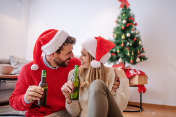 Fototapeta premium Adorable good-looking Caucasian couple with santa hats on heads sitting on floor in living room, hugging, looking at each other and holding beer. In background is christmas tree with presents.