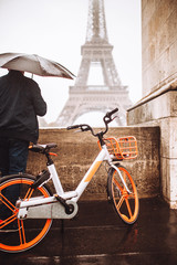 A man under an umbrella with a bicycle stopped at the bridge in the rain to look at the Eiffel Tower - a tourist cycle route