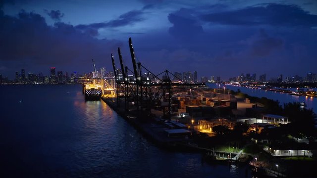 Drone flies along the docks of Port Miami. A part of the berth is lit with night lights. Two huge cargo ships are moored for unloading. The city stretches along the horizon. Lightning flashes suddenly