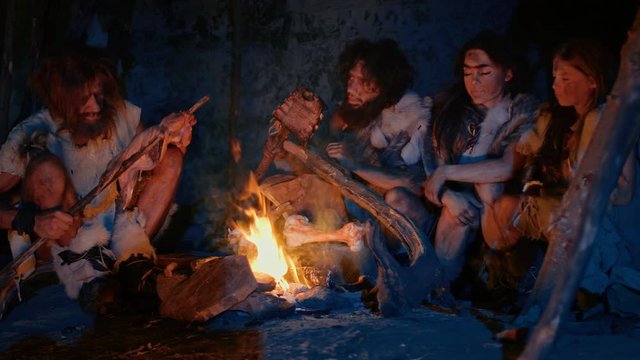 Neanderthal or Homo Sapiens Family Cooking Animal Meat over Bonfire and then Eating it. Tribe of Prehistoric Hunter-Gatherers Wearing Animal Skins Grilling and Eating Meat in Cave at Night